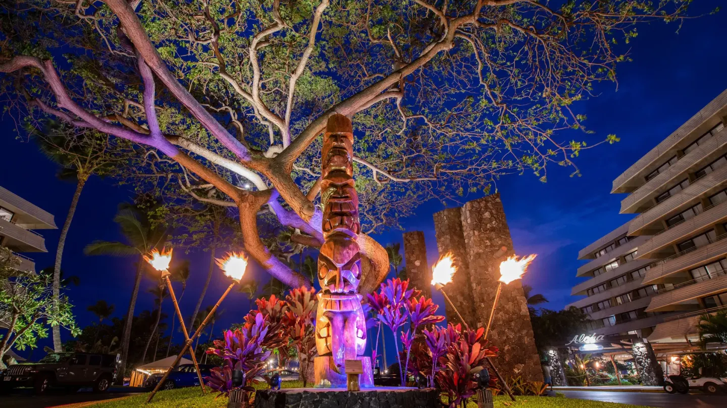 Torch-lit tiki carving and entrance gardens at night at Royal Kona Resort.