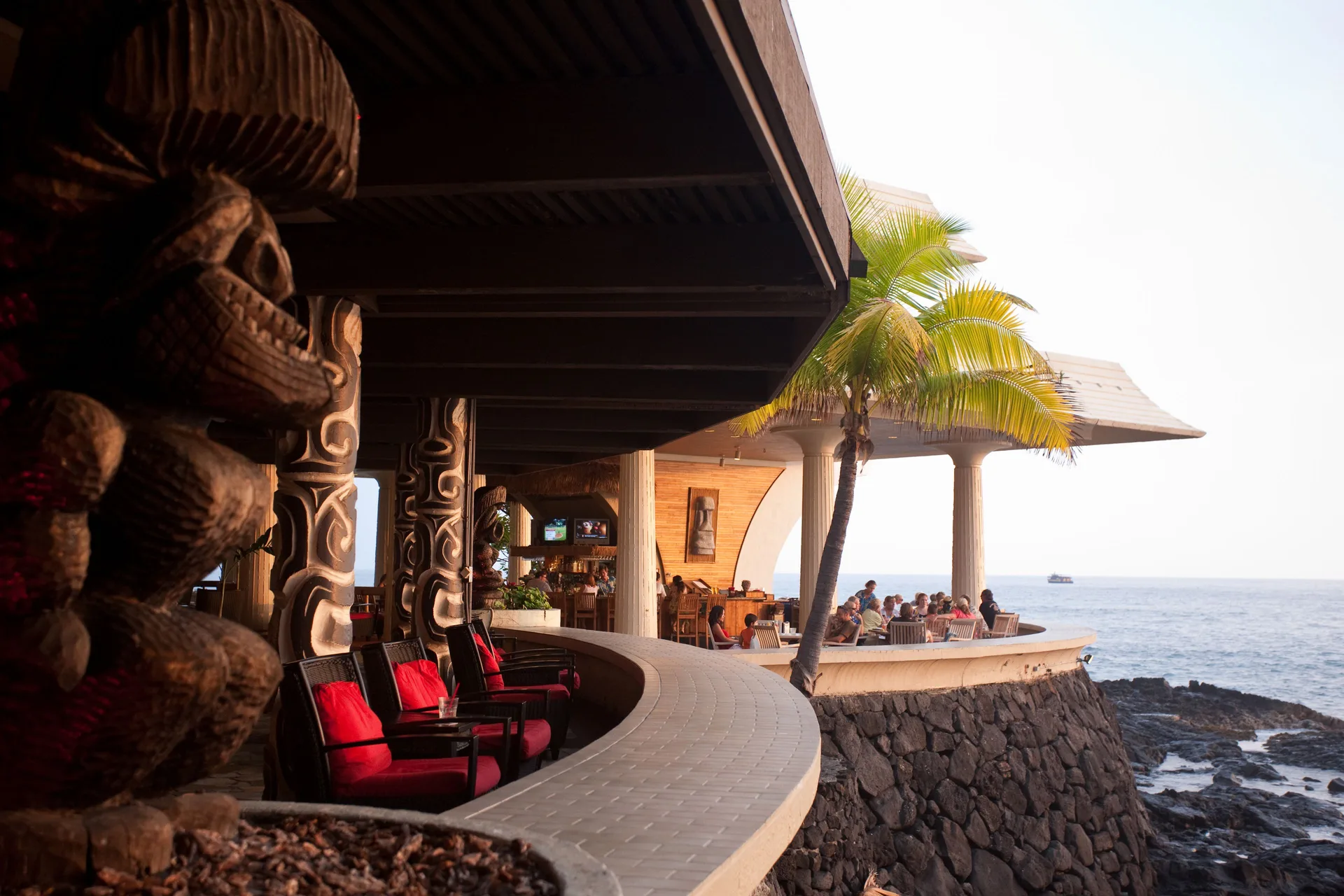Oceanfront lounge terrace with carved wood details, red chairs, palm tree, and diners at Royal Kona Resort.