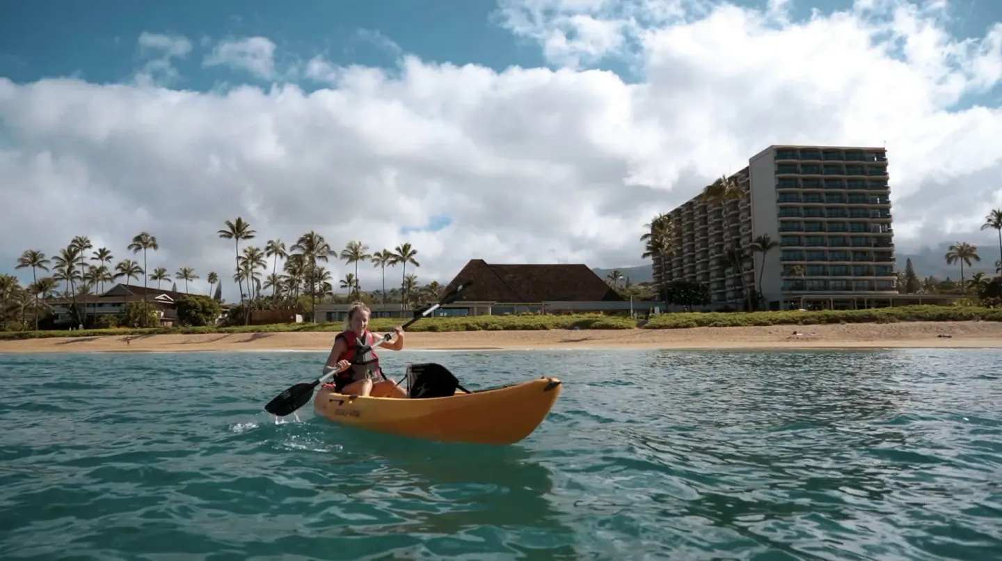 Kayaker paddling offshore with Royal Lahaina Resort & Bungalows along the beach behind.