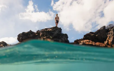 Person standing on dark ocean rocks above turquoise water at Royal Lahaina Resort & Bungalows.