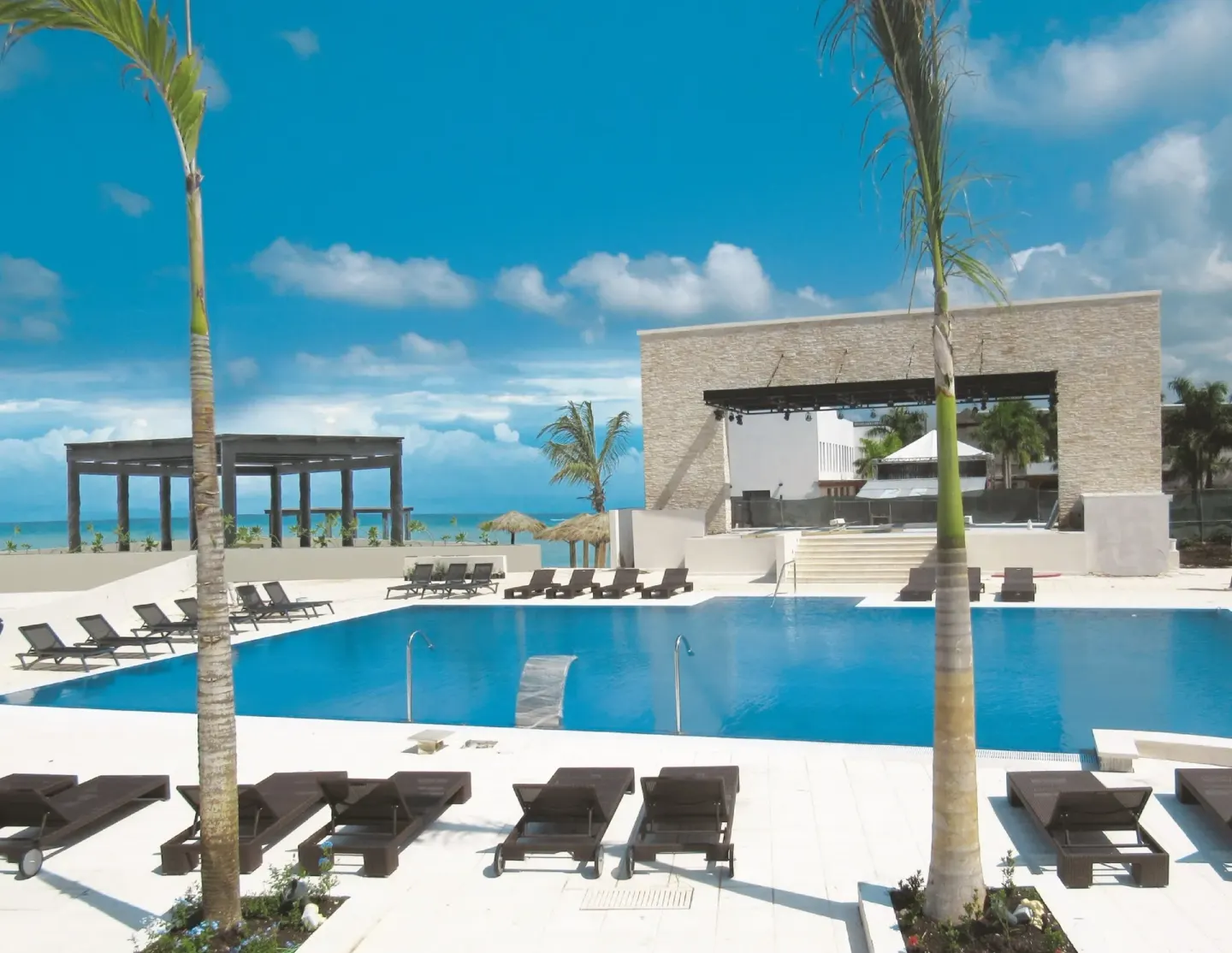 A pool deck with loungers, palms, and an ocean backdrop at Royalton Blue Waters Montego Bay.