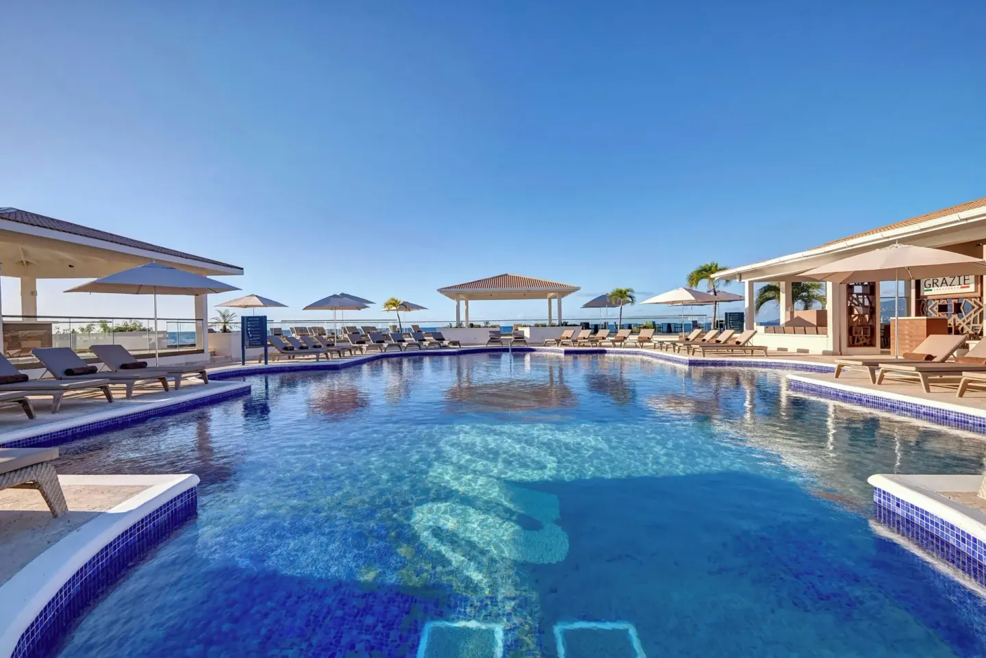 Main pool with loungers, umbrellas, ocean backdrop, and Grazie sign at Royalton Grenada.