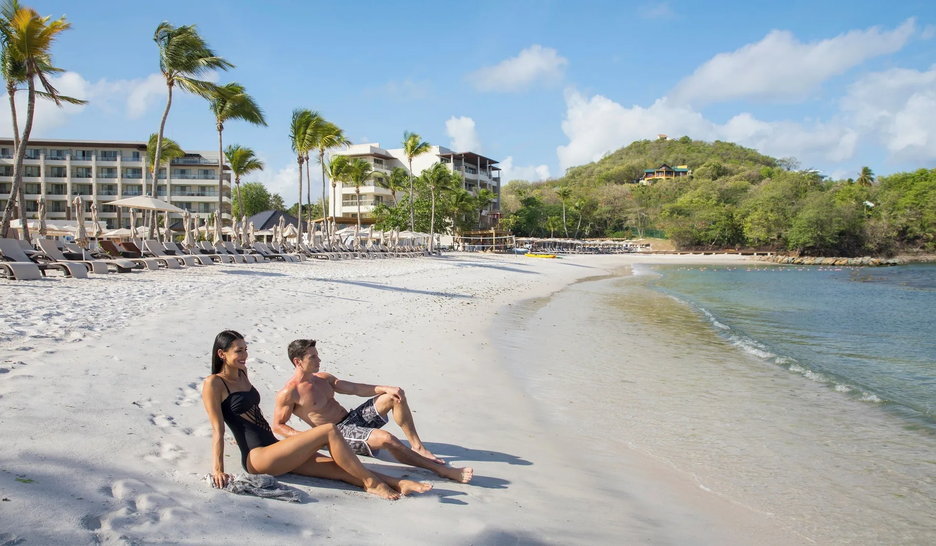 Guests sit on the beach with palm trees and resort buildings at Royalton Hideaway Saint Lucia.