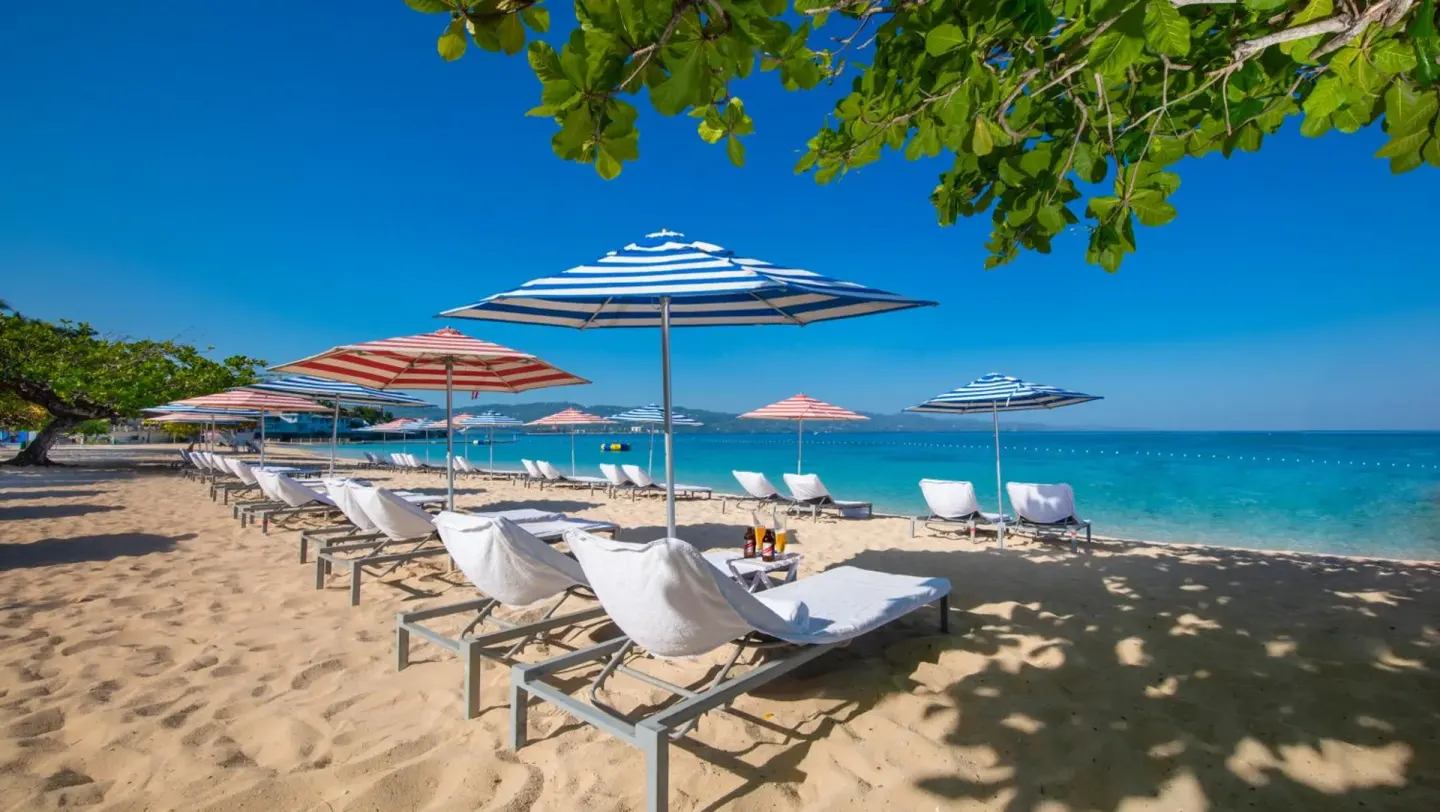 Beach loungers with striped umbrellas beside turquoise water at S Hotel Montego Bay.