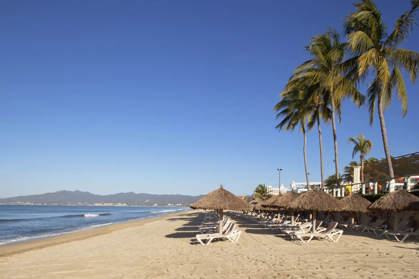 Sandy beach with palapa umbrellas, loungers, palms, ocean, and mountains at Samba Vallarta.