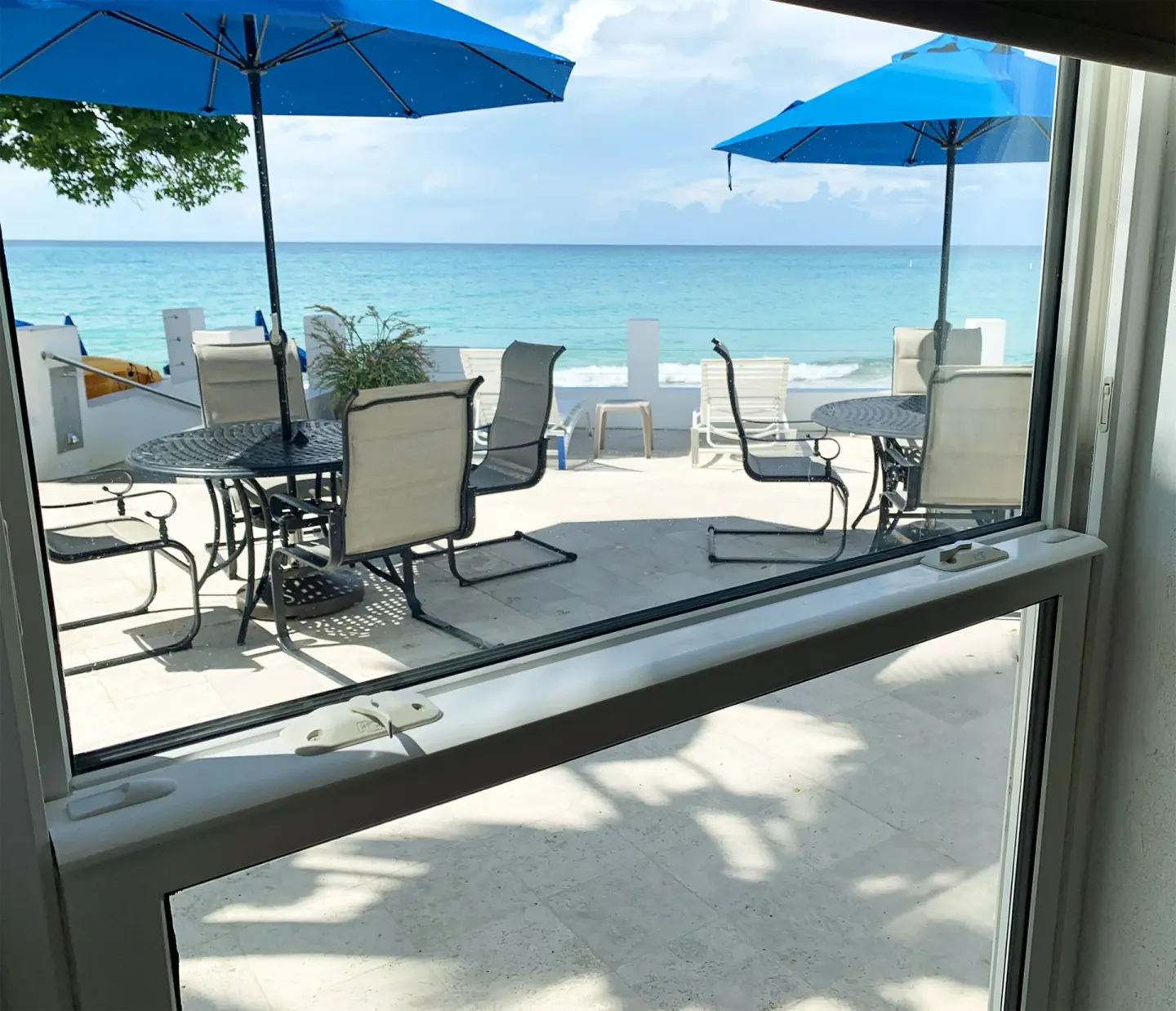 Beachfront patio tables and blue umbrellas seen through a window at Sand Castle on the Beach.