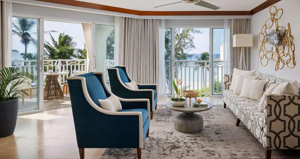 Ocean view suite living room with sofa, blue chairs, and balcony doors at Sandals Barbados.