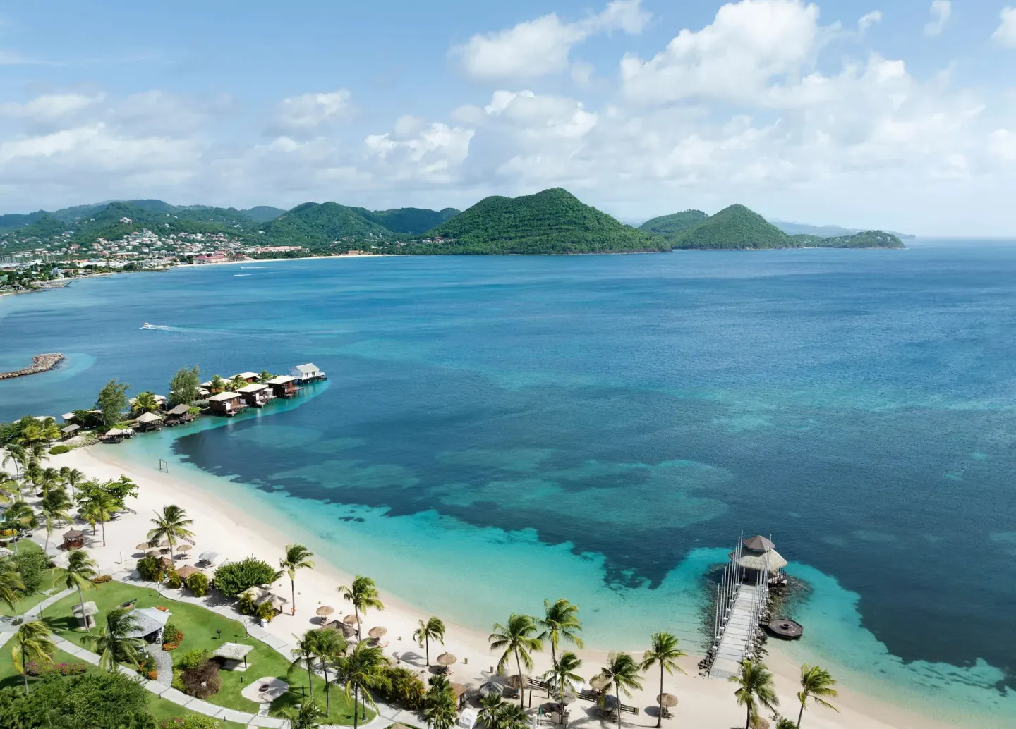 Aerial bay view of Sandals Grande St. Lucian with beach, pier, palms, and surrounding hills.