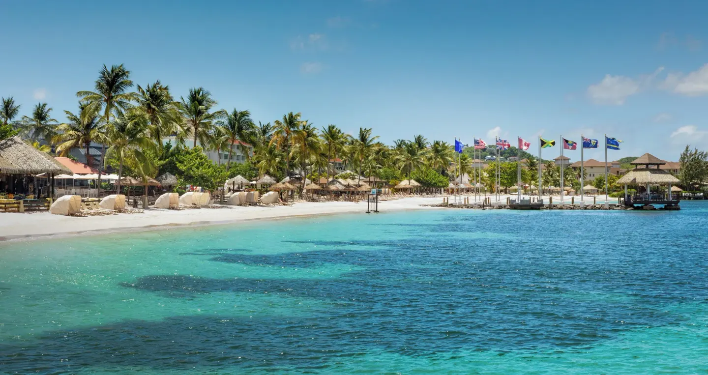 Beachfront at Sandals Grande St. Lucian with palm trees, flags, thatched shades, and blue water.