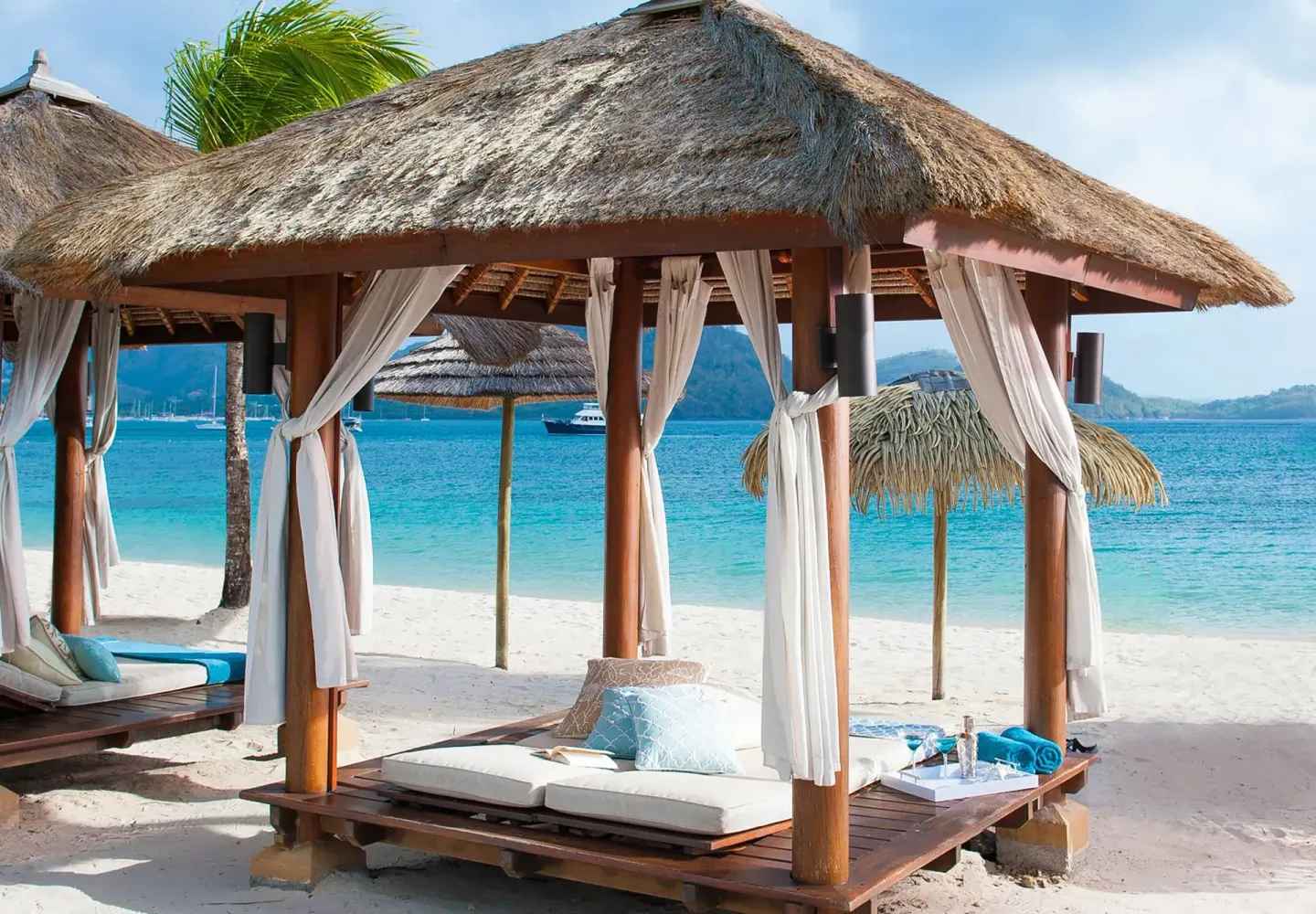 Thatched beach cabanas at Sandals Grande St. Lucian with cushioned loungers beside turquoise water.