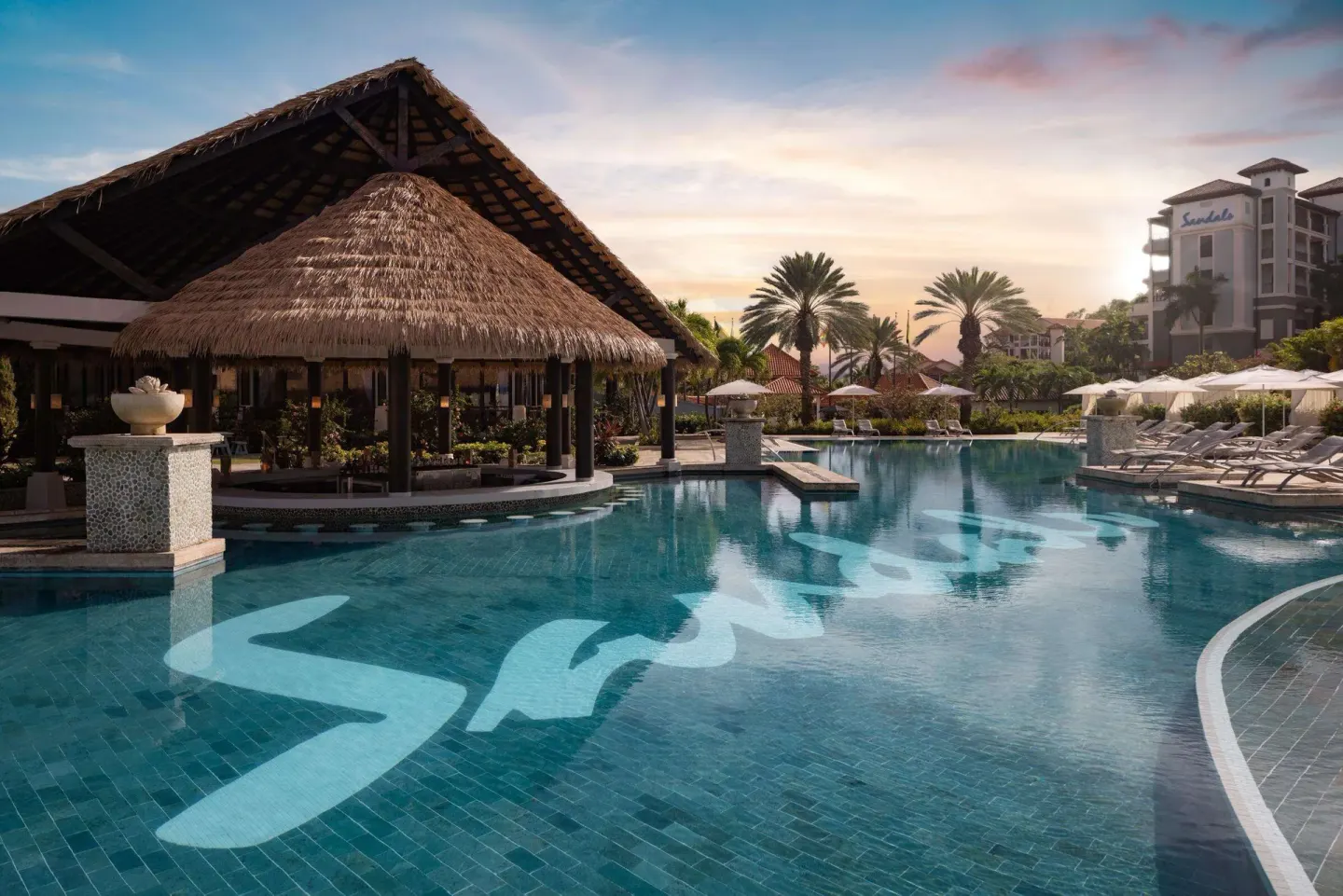 Resort pool at sunset with thatched pavilion and palm trees at Sandals Grenada.