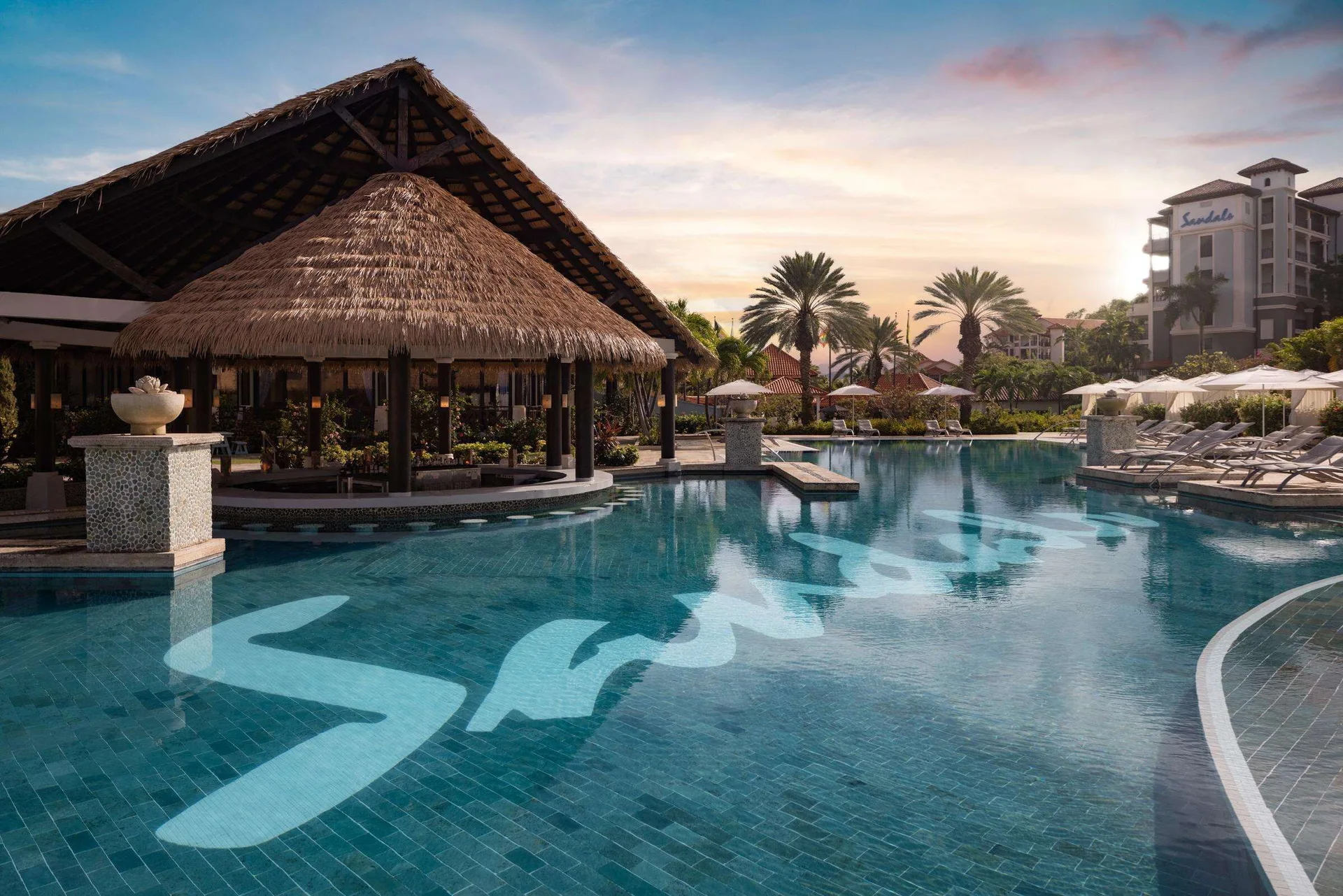 Resort pool at sunset with thatched pavilion and palm trees at Sandals Grenada.