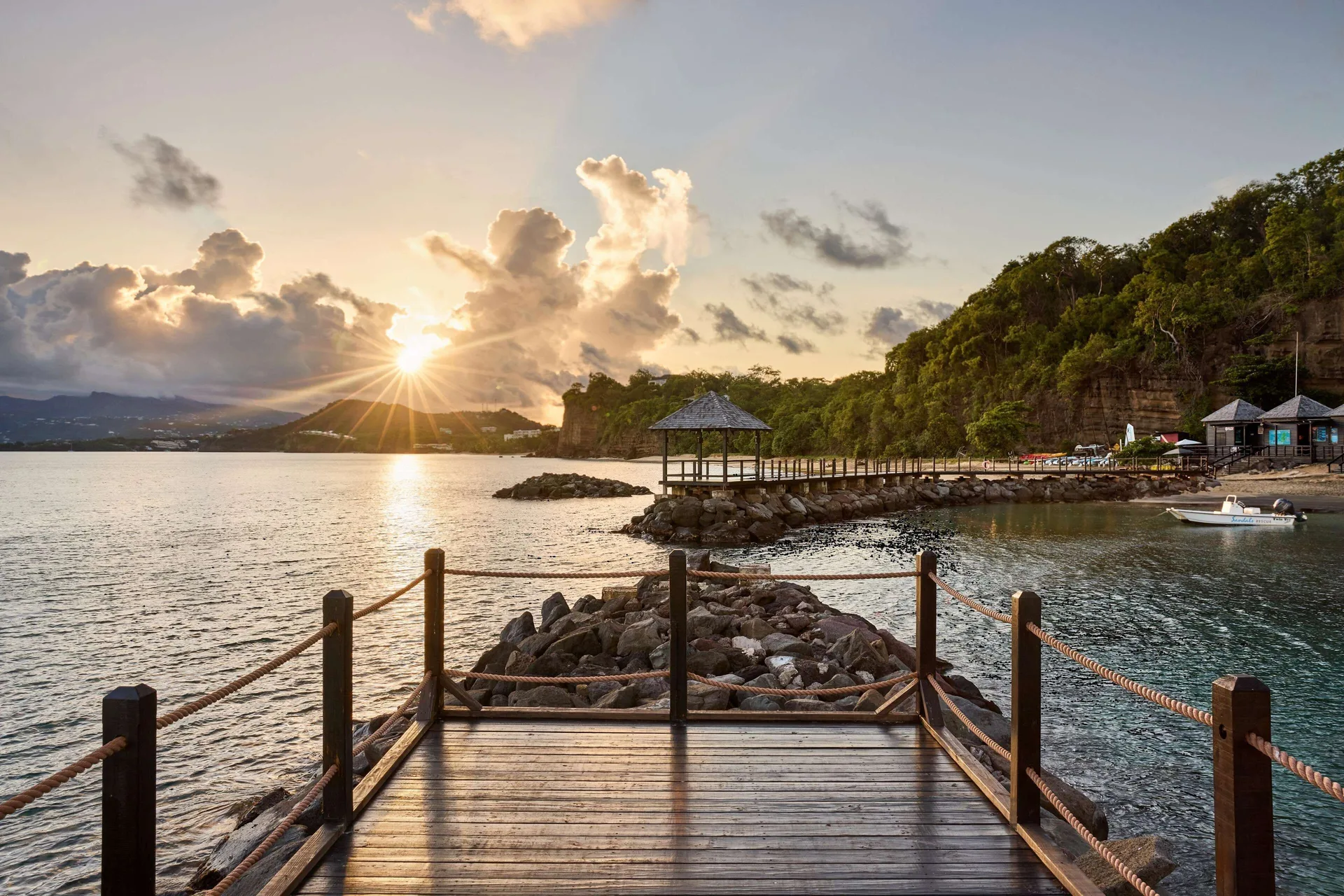Sunset shines over a wooden deck, rocky shoreline, and calm bay at Sandals Grenada.