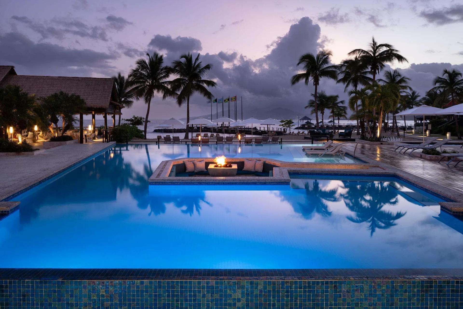 Twilight pool with fire pit seating, palms, and beach beyond at Sandals Grenada.