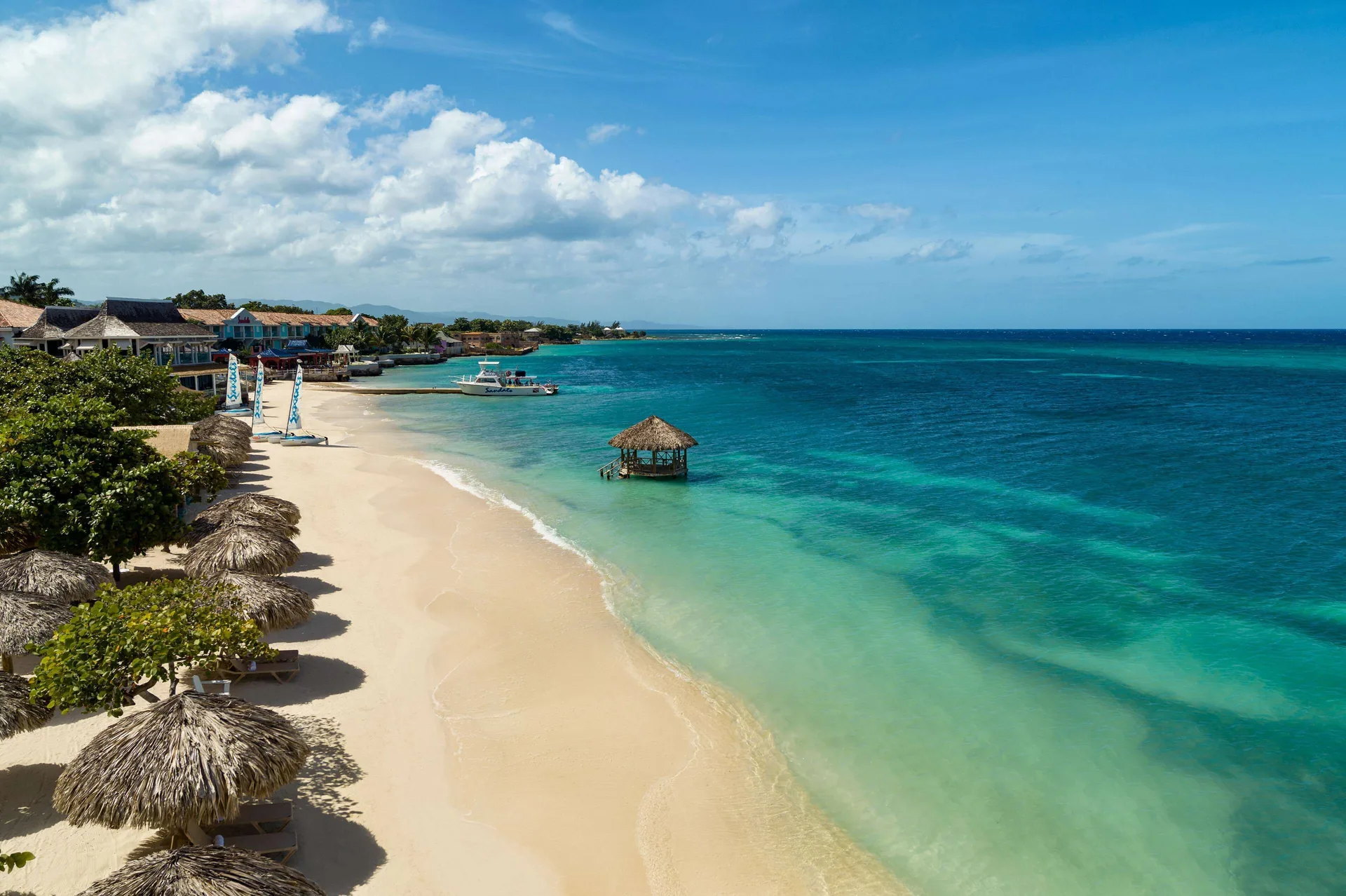 Wide beach view with palapas, a small boat, and turquoise sea at Sandals Montego Bay.