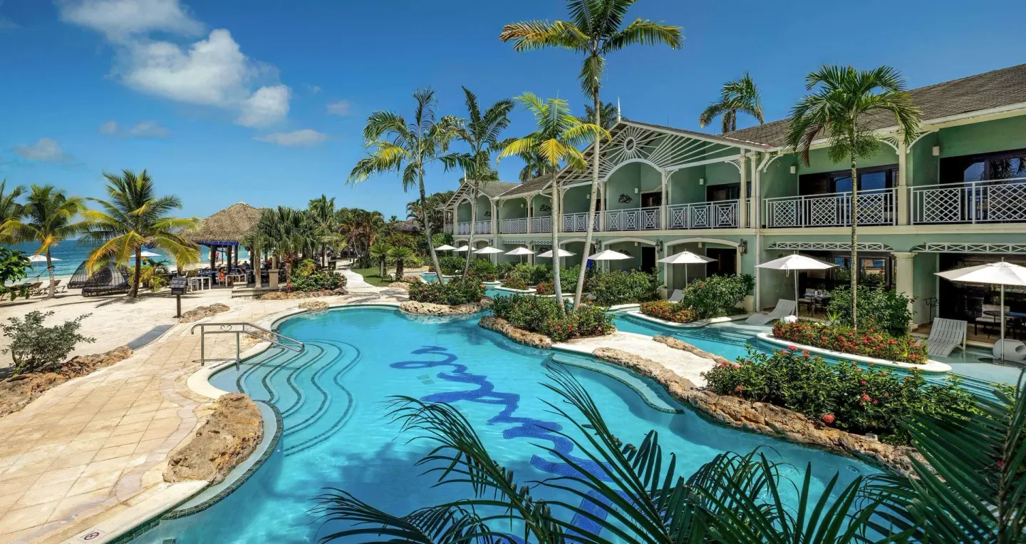 Beachside pool beside a green guest building, palms, loungers, and sand at Sandals Negril.