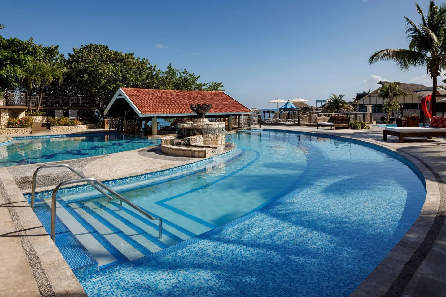 Curved pool with steps, a stone fountain, and a red-roof pavilion at Sandals Ochi.