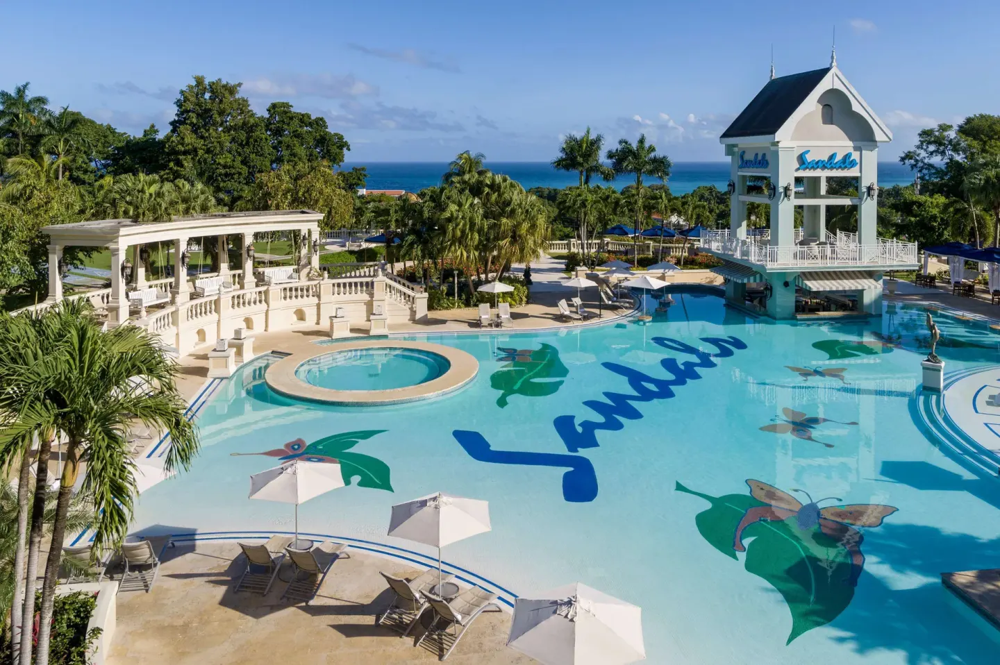 Main pool with a tower building, umbrellas, palms, and the sea beyond at Sandals Ochi.