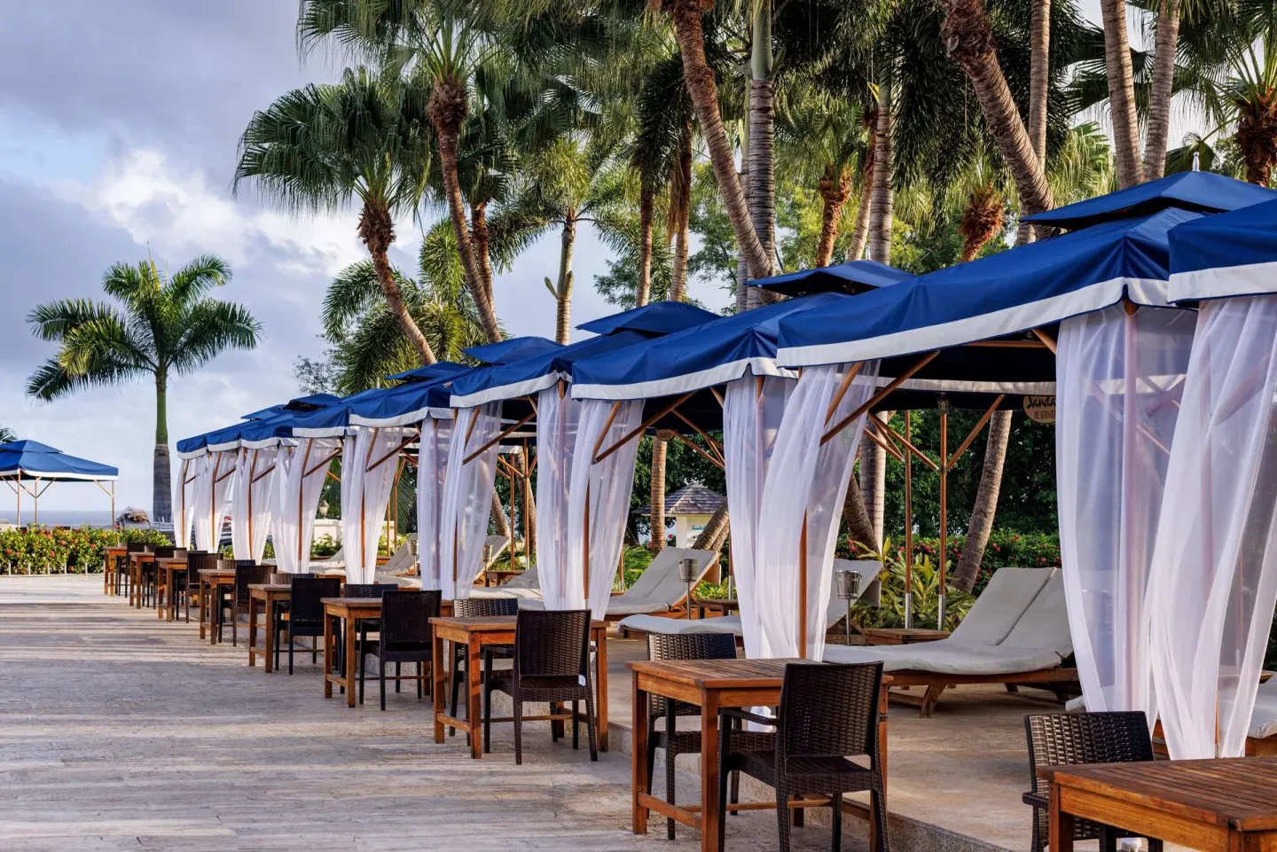 Blue-canopied cabanas with lounge chairs line a palm-shaded deck at Sandals Ochi.