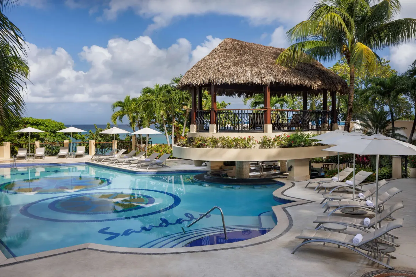 Pool with a thatched pavilion, loungers, umbrellas, and ocean glimpses at Sandals Ochi.
