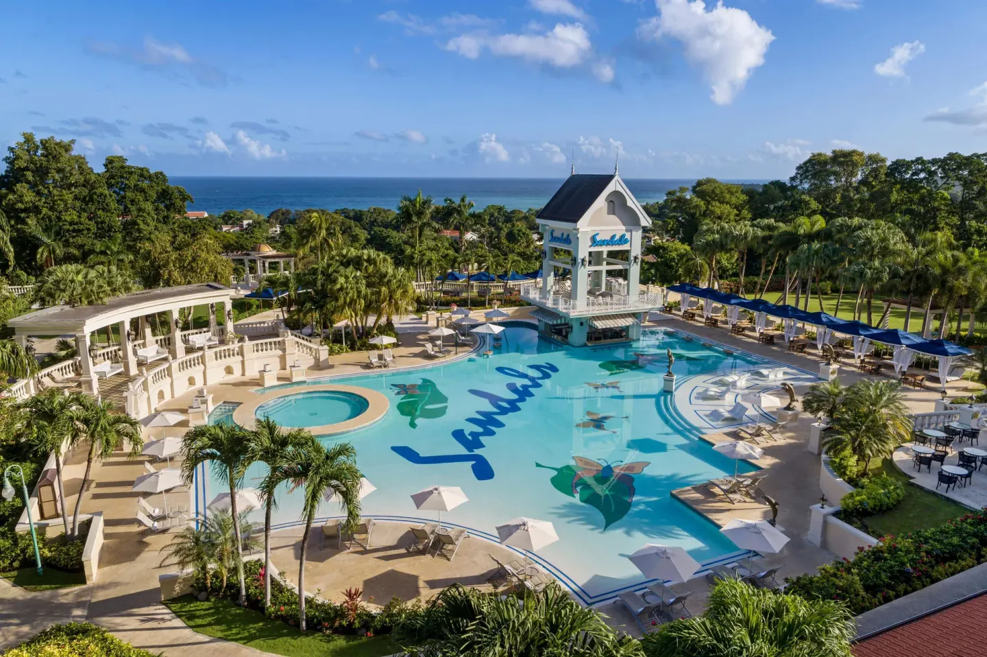 Wide aerial view of the main pool, palms, cabanas, and ocean beyond at Sandals Ochi.
