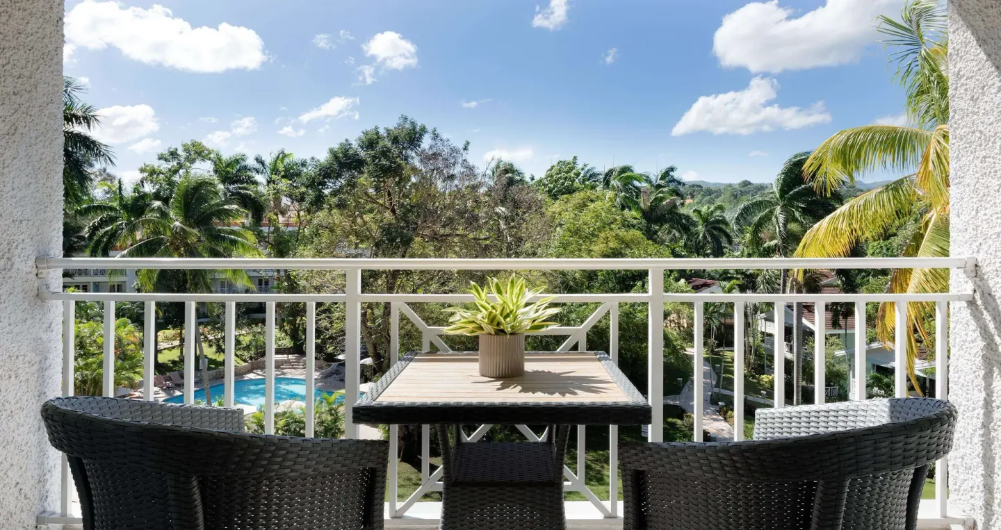 Furnished garden-view balcony at Sandals Ochi with two chairs, a table, palms, and pool below.