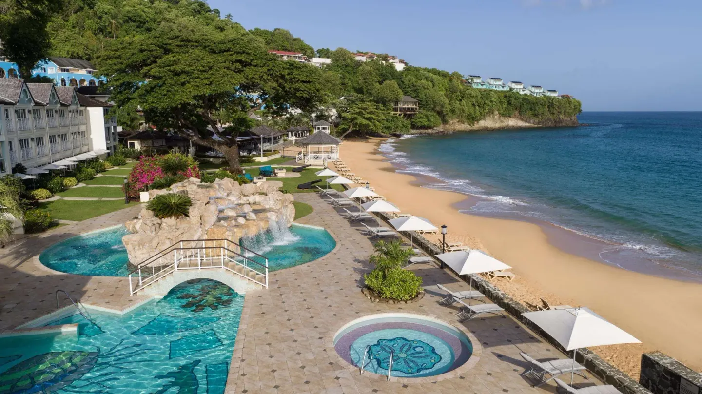 Pool, beach, and sea at Sandals Regency La Toc with loungers and hillside greenery.
