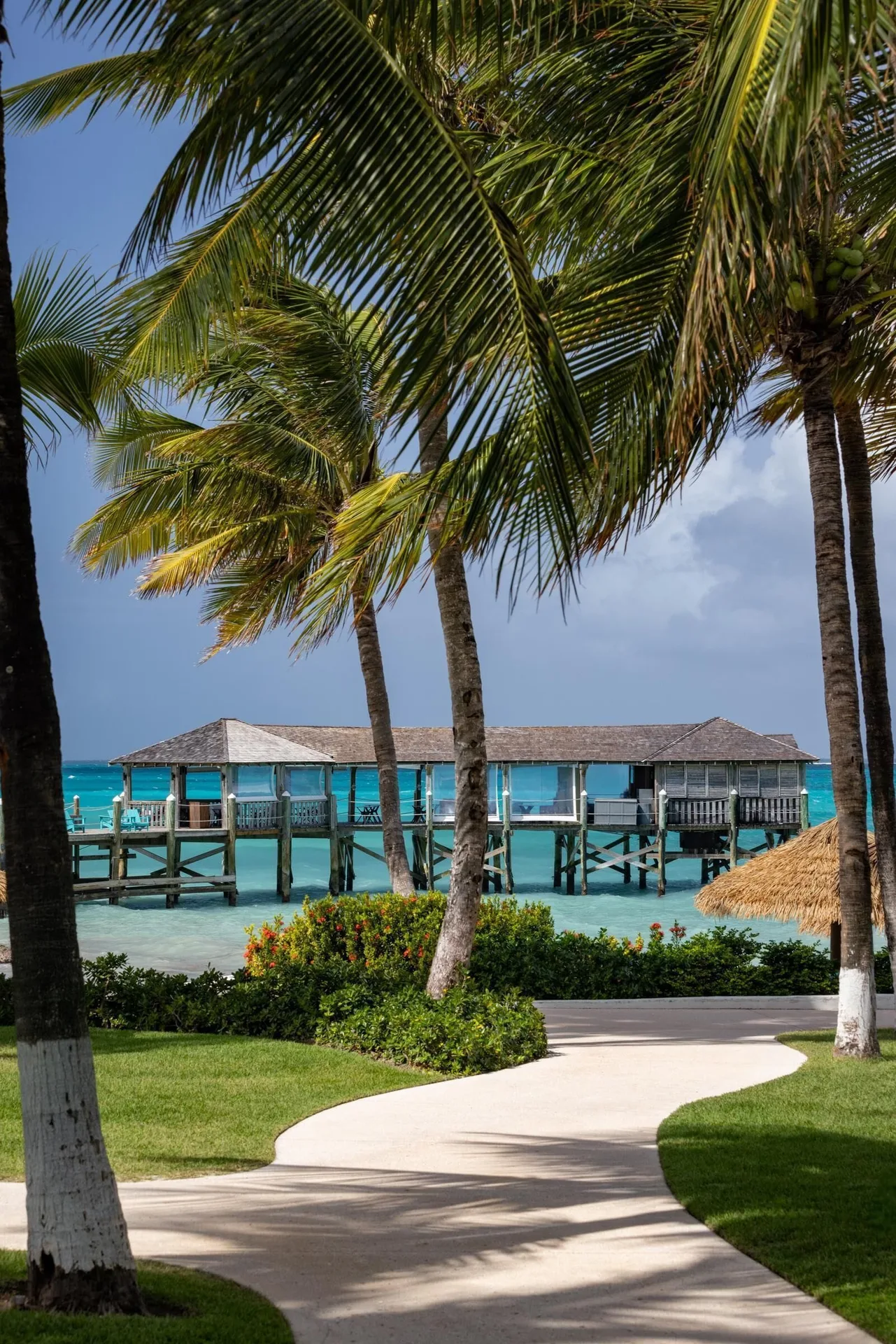 Palm-lined oceanfront walkway near Sandals Royal Bahamian with turquoise water and pier buildings.