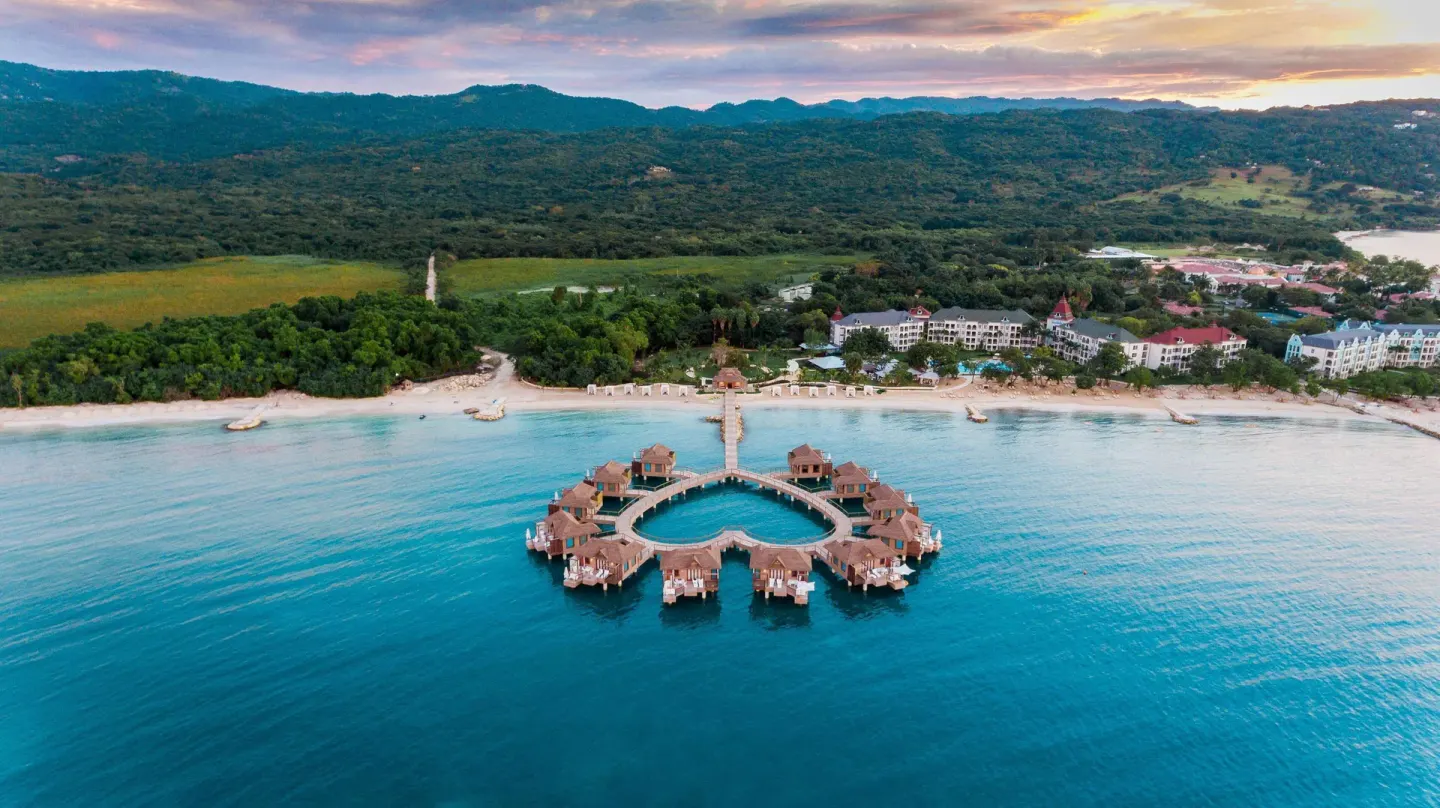 Aerial view of overwater villas, beach, and hills at Sandals South Coast at dusk.