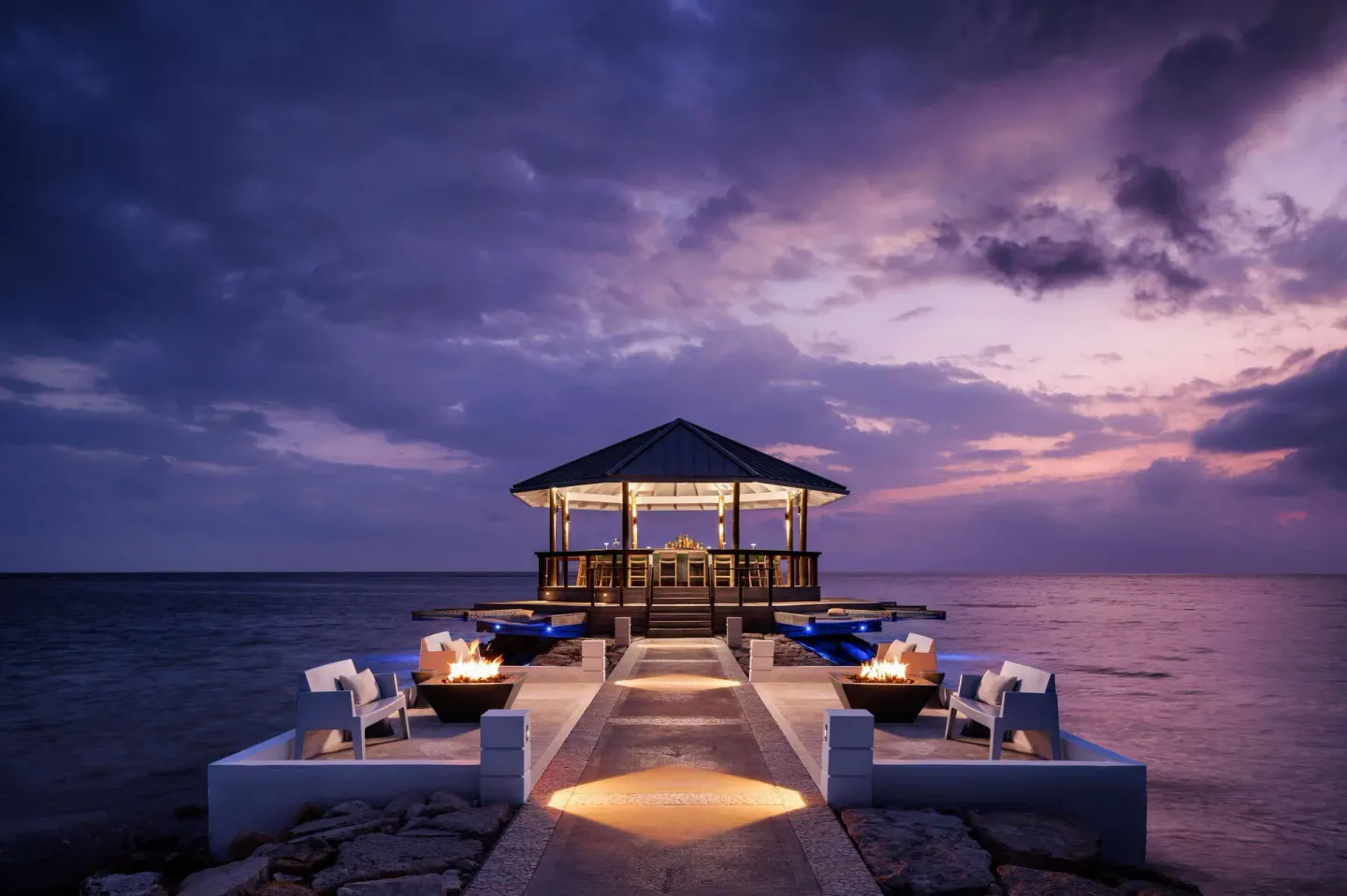 Overwater deck with fire bowls and a lit pavilion at Sandals South Coast at dusk.