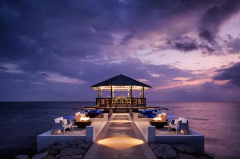 Overwater deck with fire bowls and a lit pavilion at Sandals South Coast at dusk.