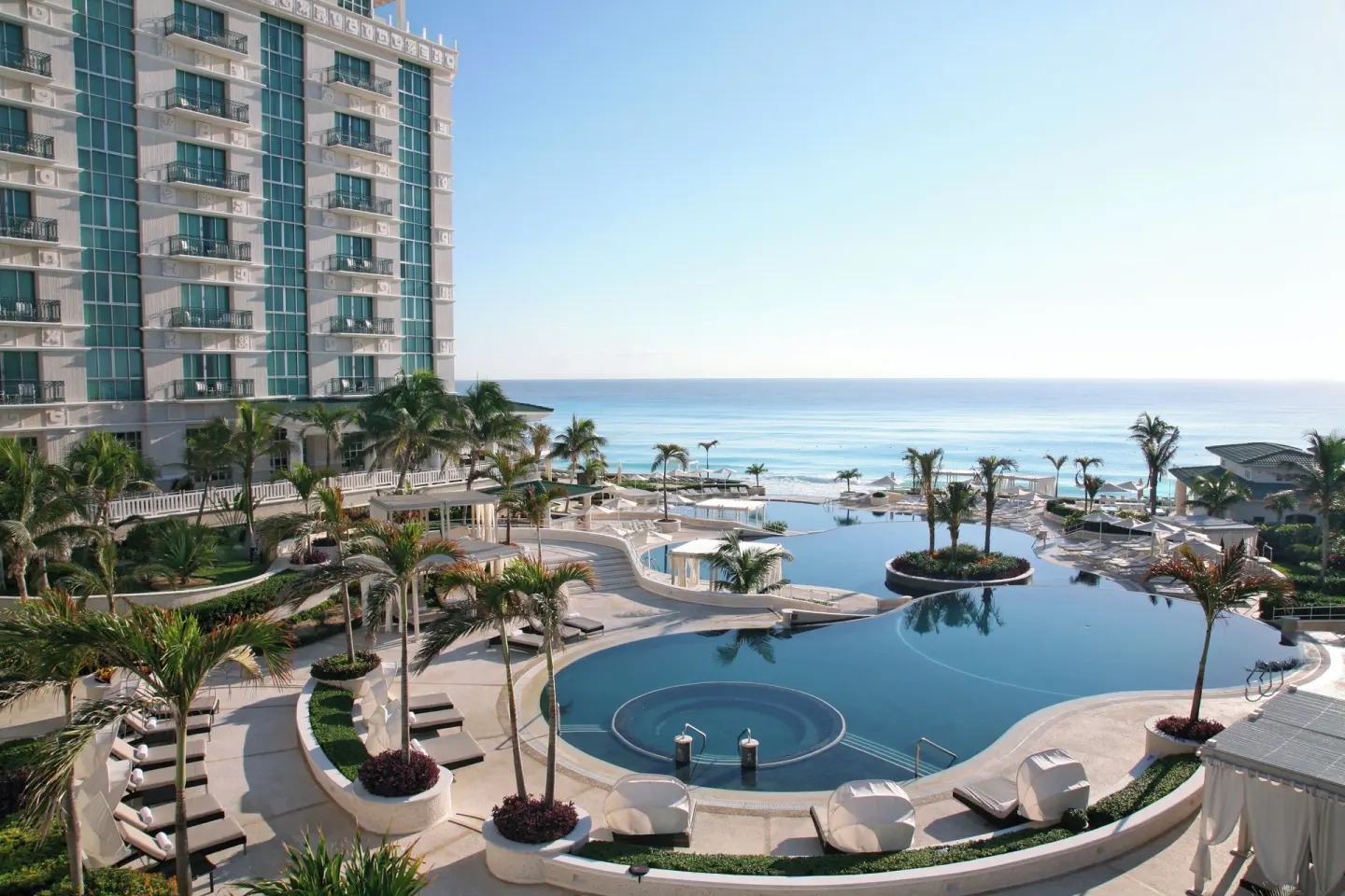 Oceanfront pools, palms, loungers, and the resort tower at Sandos Cancún.