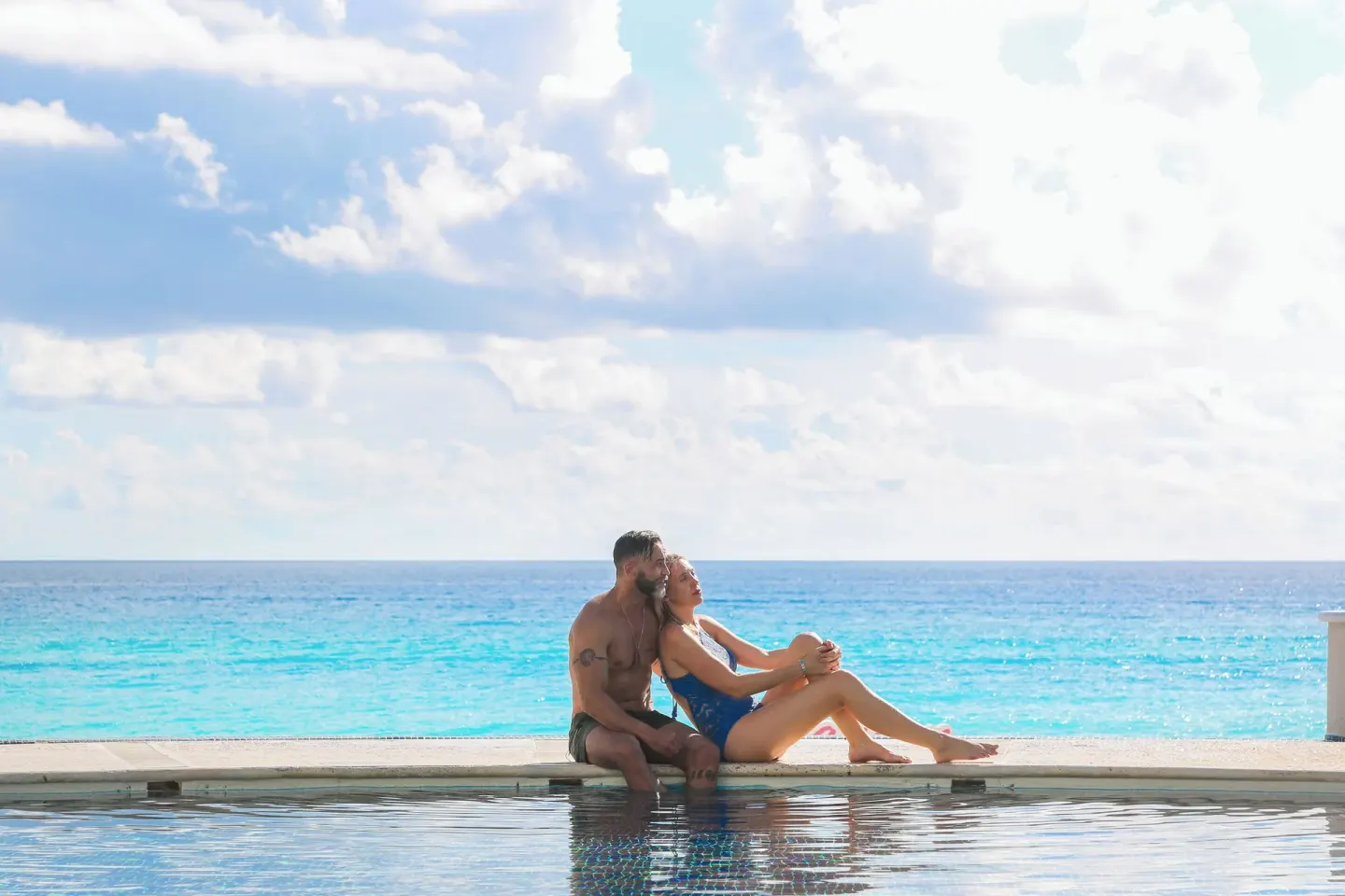 A couple sits at the pool edge with the turquoise ocean behind them at Sandos Cancún.