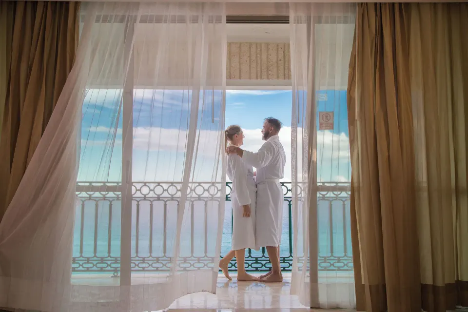 Couple in robes on the Caribe Suite balcony behind sheer curtains at Sandos Cancún.
