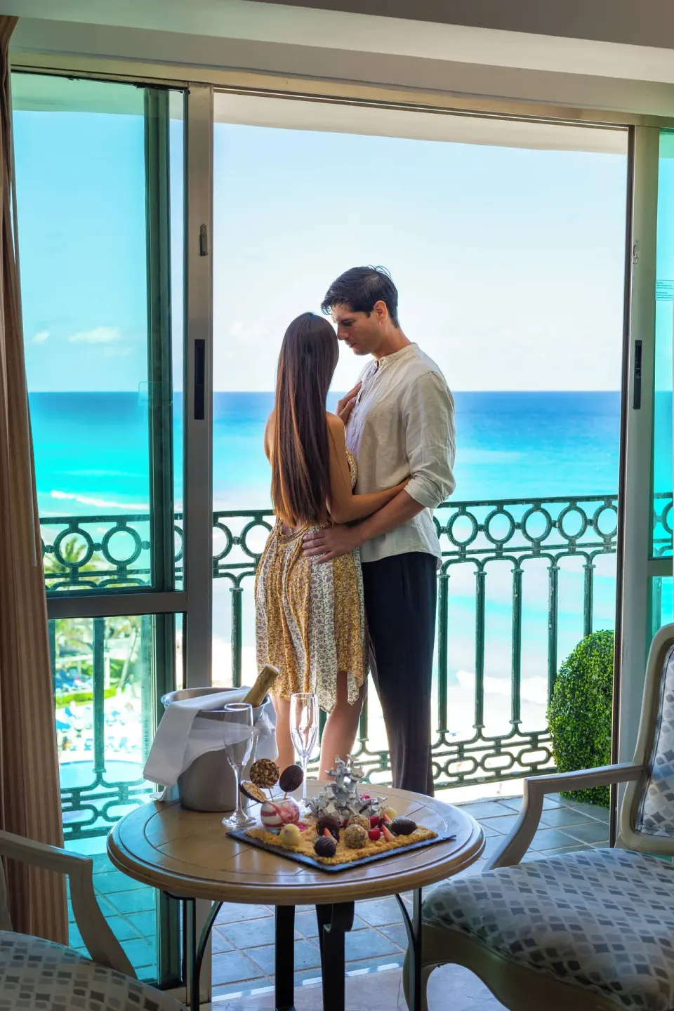 Couple on a Caribe Suite balcony with ocean view and welcome tray at Sandos Cancún.