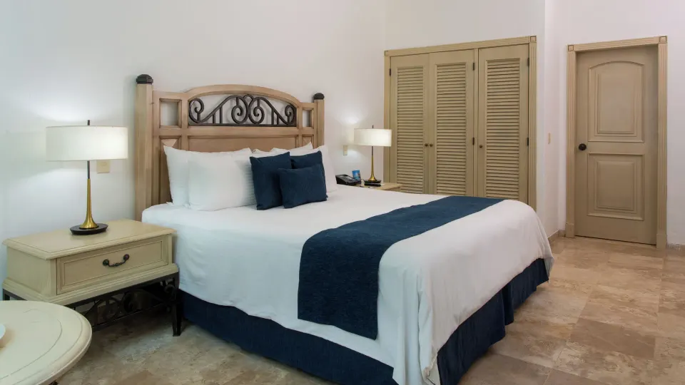 Sandos Cancún bedroom with a king bed, blue accents, lamps, closet doors, and entry door at Sandos Cancún.