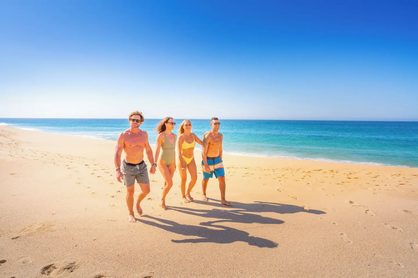 Four guests walk along the sunny beach beside turquoise water at Sandos Finisterra.