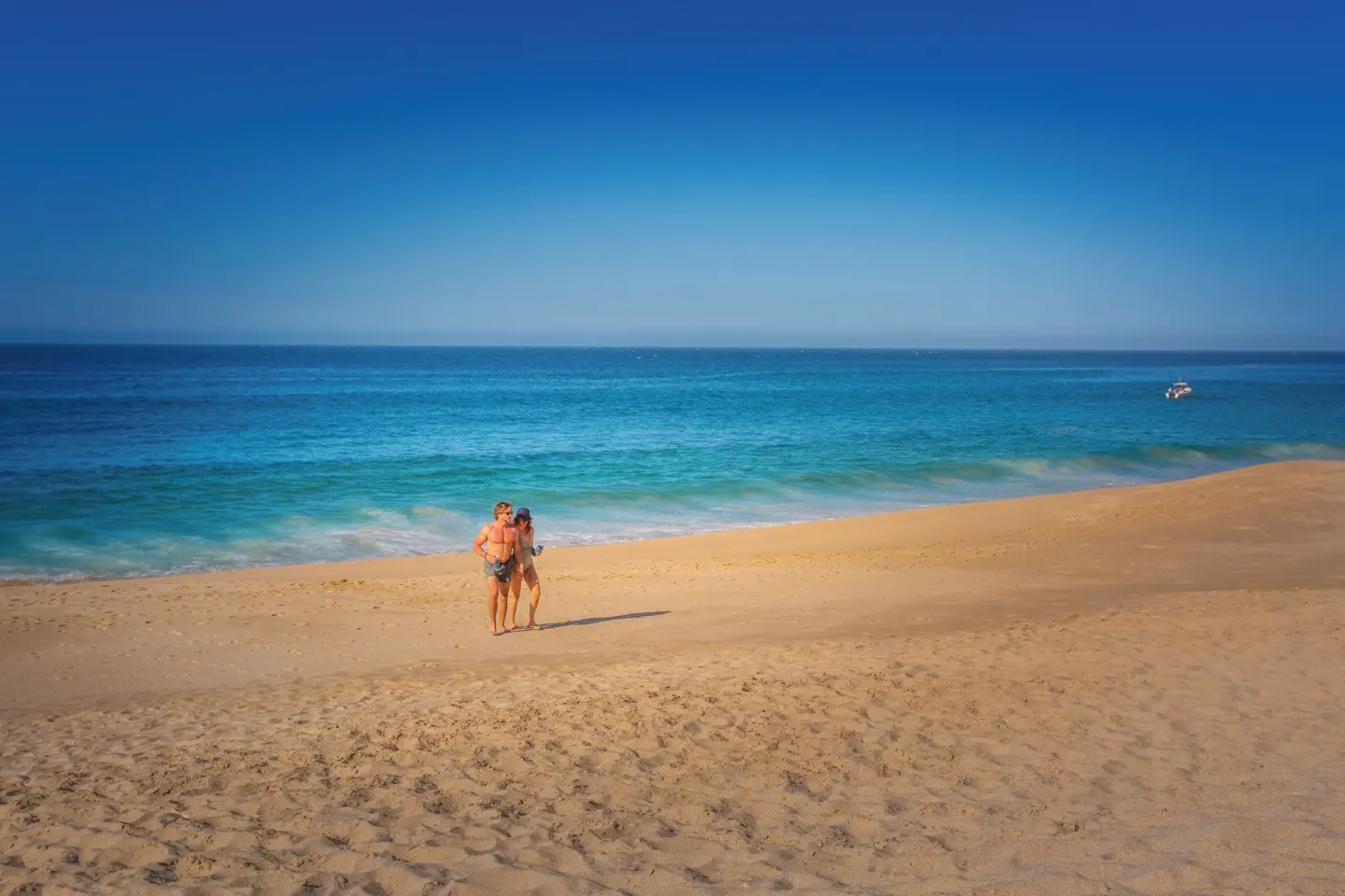 A couple stands on a wide sandy beach with ocean and a small boat at Sandos Finisterra.