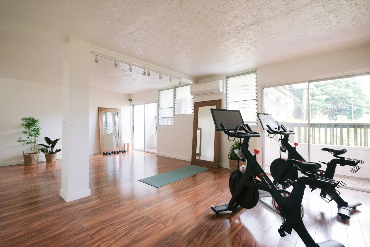 A bright fitness room with bikes, yoga mat, mirrors, and wood floors at SCP Hilo Hotel.