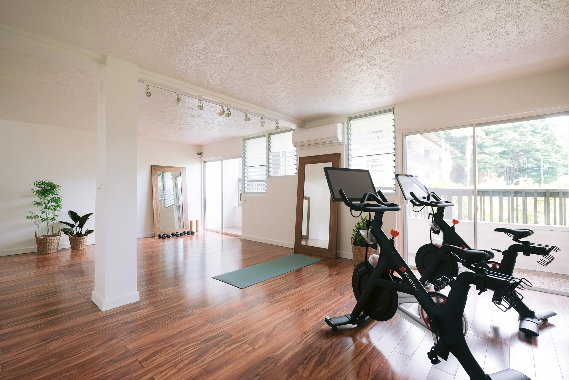 A bright fitness room with bikes, yoga mat, mirrors, and wood floors at SCP Hilo Hotel.