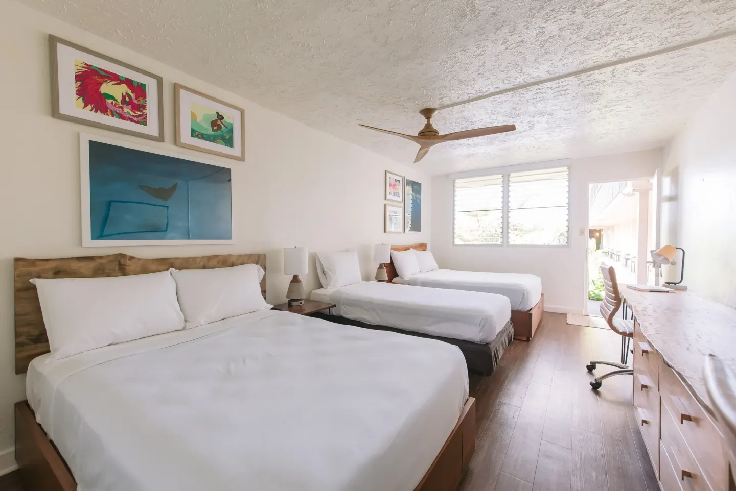 Three-bed guest room with dresser, desk, and bright windows at SCP Hilo Hotel.