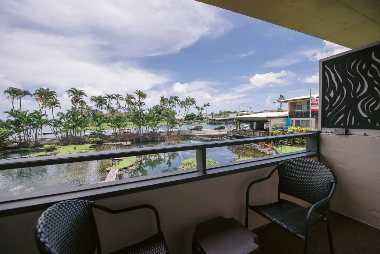 Room balcony with chairs overlooking ponds, palms, and Hilo Bay at SCP Hilo Hotel.