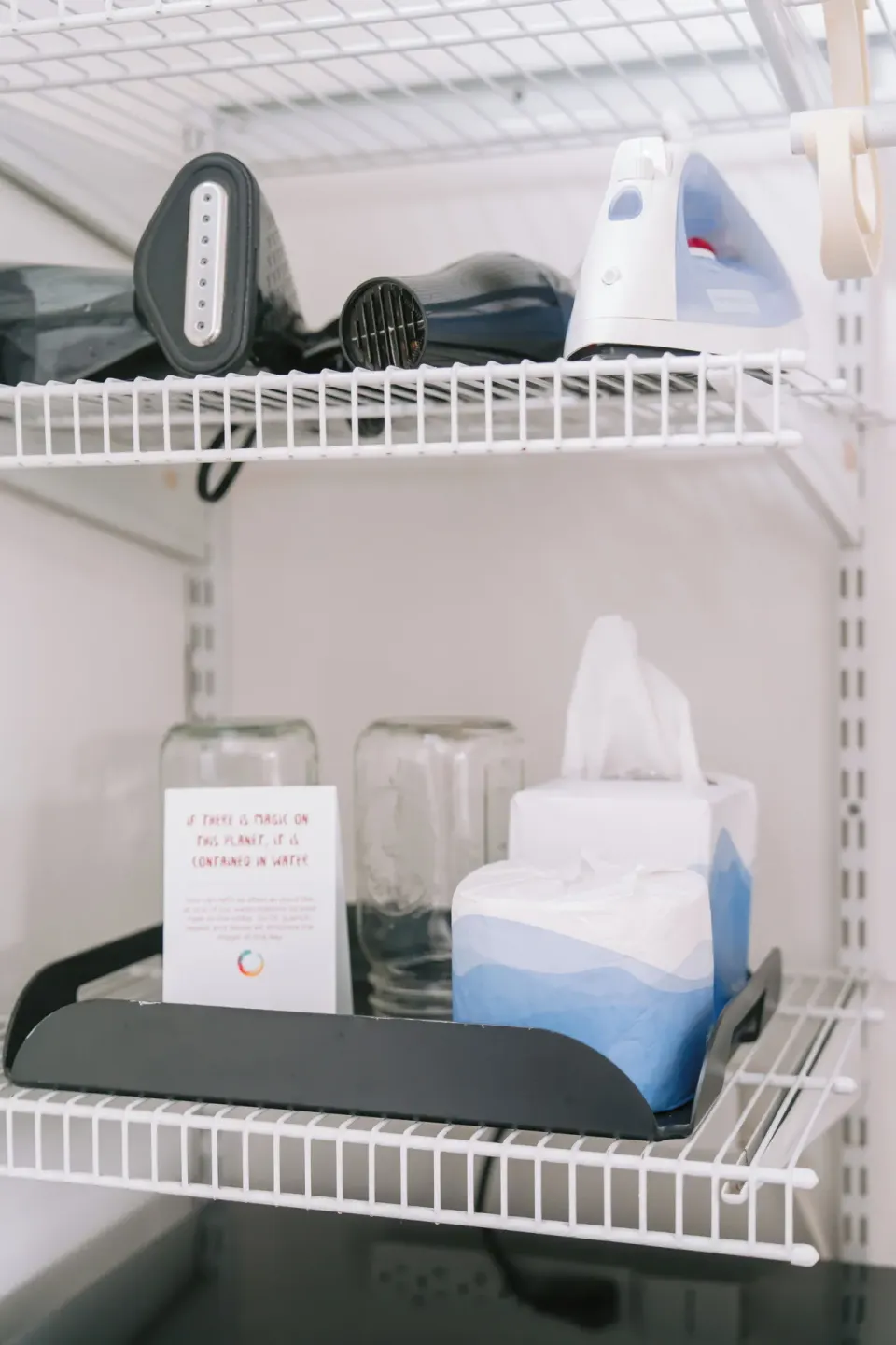 Closet shelf with iron, hair dryer, glasses, and tissues in a guest room at SCP Hilo Hotel.
