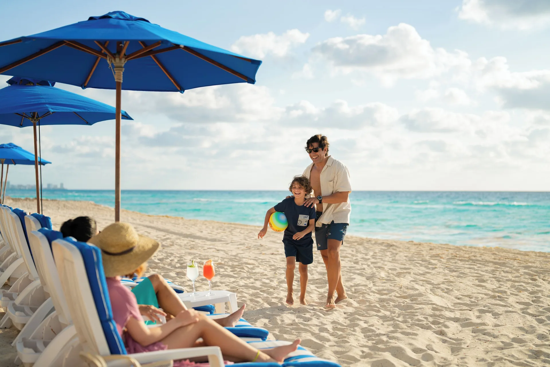A family walks past beach loungers and blue umbrellas at Seadust Cancún Family Resort.