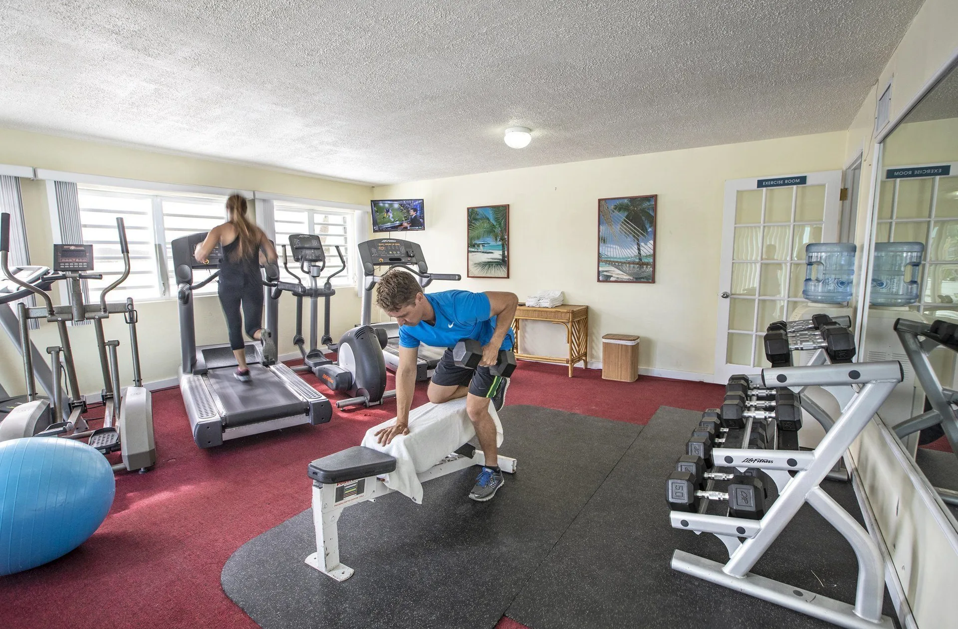 Guests exercise in the fitness room at Secret Harbour Beach Resort with treadmills and free weights visible.