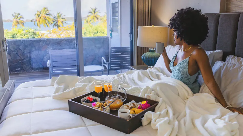 Guest relaxes in bed with breakfast and balcony view at Secret Harbour Beach Resort.