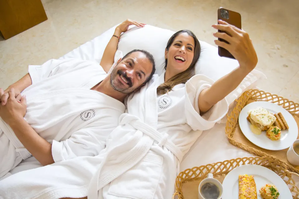 Couple in robes taking a selfie over breakfast in bed at Sheraton Buganvilias Puerto Vallarta.