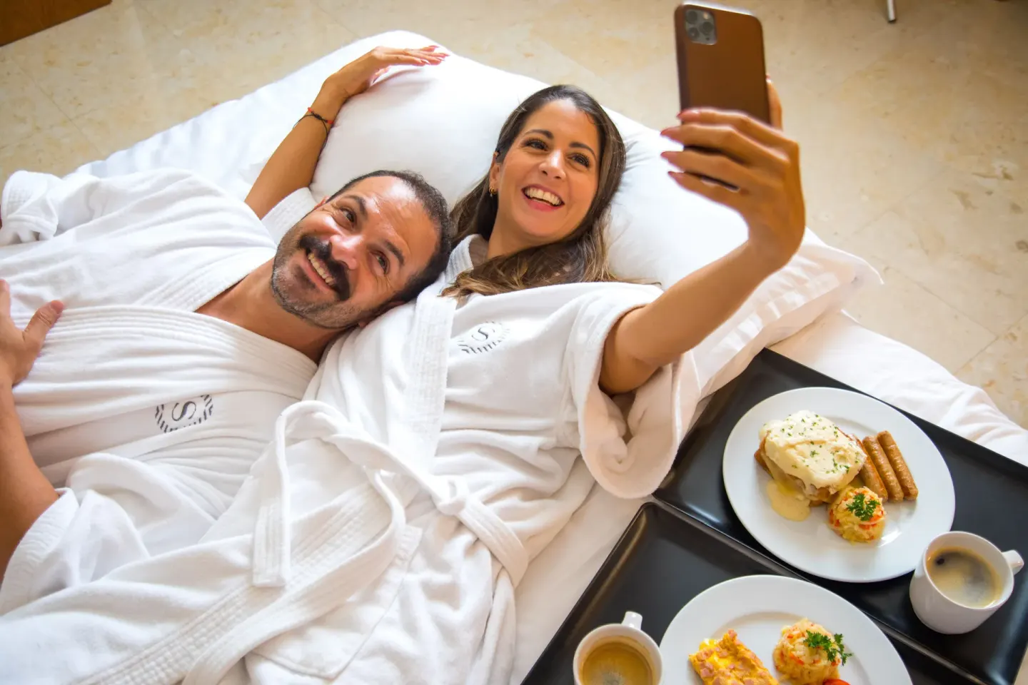 Couple in robes smiling with breakfast trays on the bed at Sheraton Buganvilias Puerto Vallarta.