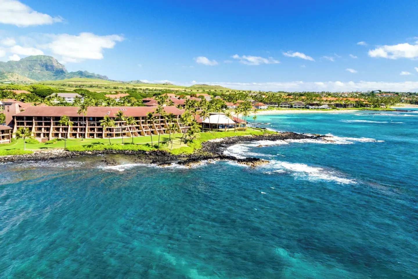 Aerial coastline view of Sheraton Kauai Resort with blue ocean, lava rocks, and distant hills.