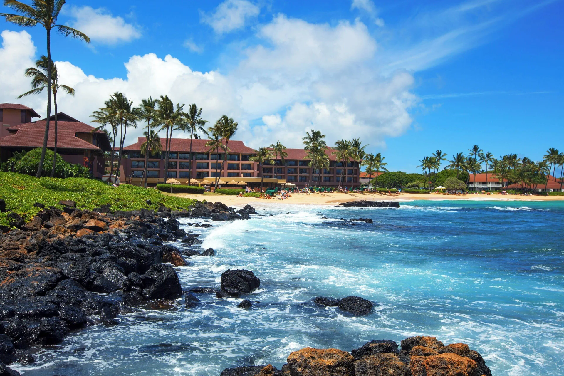 Beachfront buildings at Sheraton Kauai Resort overlook turquoise water and black lava rocks.